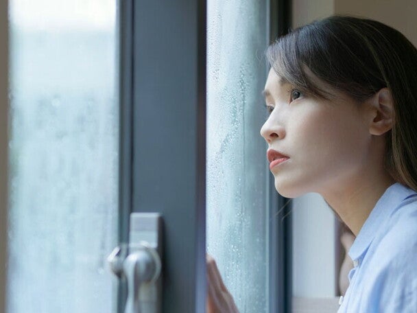 A young Asian woman, looking out at the rain, wondering what typhoon safety precautions to take for her family.