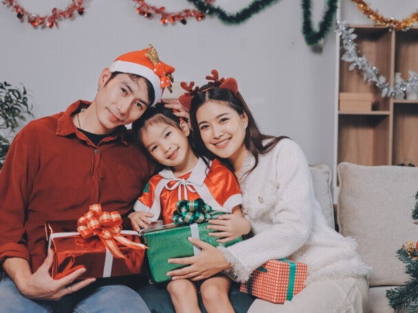 A young Asian couple with their little girl posing for a Christmas photoshoot while holding Christmas presents and wearing holiday headwear.