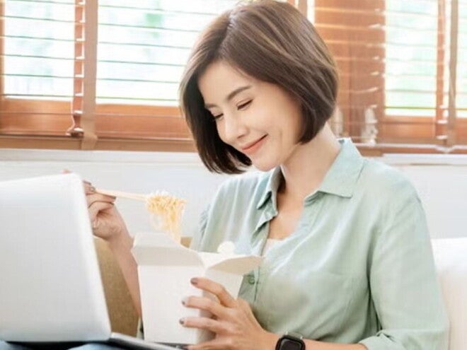 Young Filipina experiences a food craving and enjoys takeout while sitting on the sofa with her laptop on her lap.