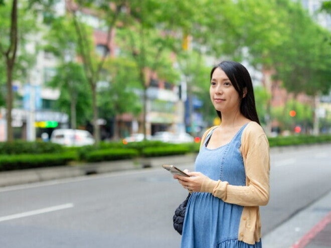 A 17-week-pregnant Asian woman holding her smartphone while standing on the street.