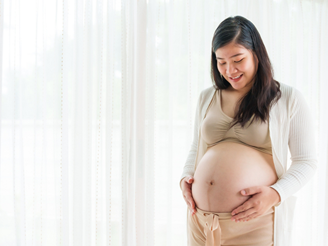A 31 weeks pregnant Asian woman standing by the window, holding her large belly bump