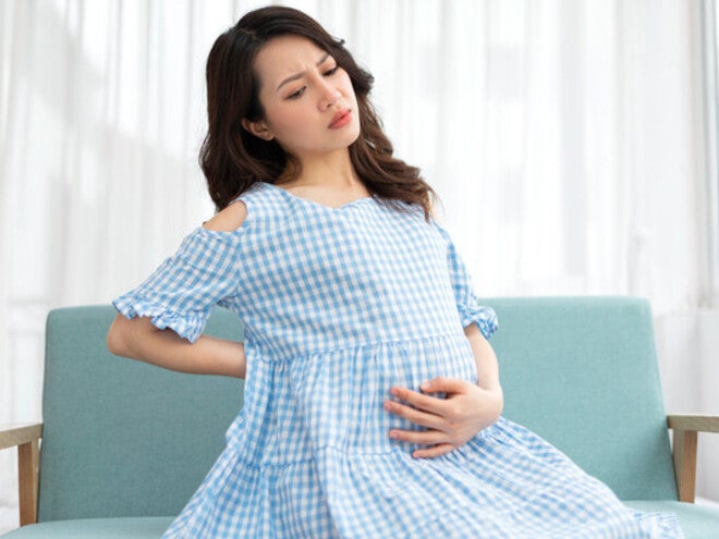 A pregnant Asian woman is sitting on the sofa and holding her belly in discomfort due to cramps during pregnancy