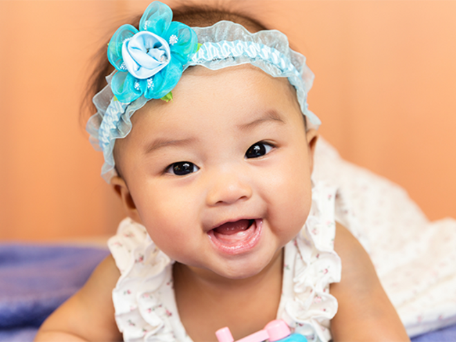 A smiling Asian little girl on her tummy, showing off baby’s first tooth