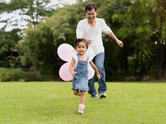 A smiling Asian preschooler girl running in the park with her dad for a budget-friendly day out. 