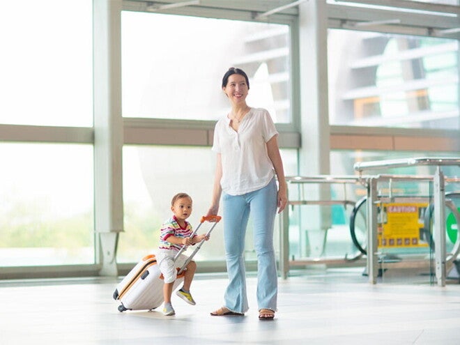 A young Asian mom with her young toddler riding on her luggage at the airport.