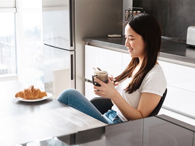 A young Asian woman is sitting in the kitchen drinking coffee during pregnancy