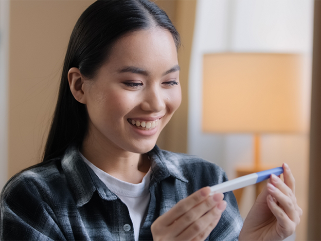 A young Asian woman smiling as she looks at a pregnancy test and curious about the stages of pregnancy.
