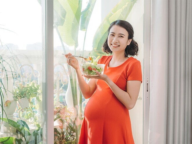 A young Asian woman standing by the window and holding a bowl of salad as part of her healthy pregnancy meal plan. A young Asian woman standing by the window and holding a bowl of salad as part of her healthy pregnancy meal plan.