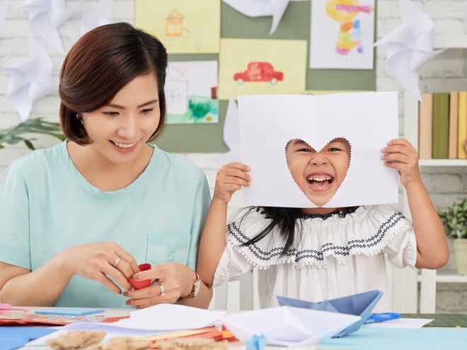 A young girl showing a heart cutout on paper, doing arts and craft for kids at home with her mom.