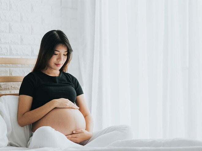 A young pregnant Asian woman holding her belly and seated in bed, alert to the signs of labor A young pregnant Asian woman holding her belly and seated in bed, alert to the signs of labor