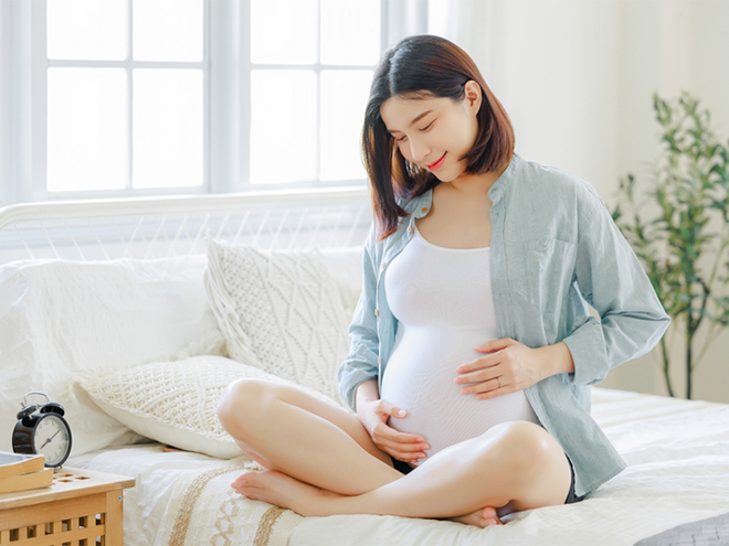 A young, pregnant Asian woman sitting cross-legged on the bed, touching her belly, aware of the importance of recognizing preeclampsia symptoms A young, pregnant Asian woman sitting cross-legged on the bed, touching her belly, aware of the importance of recognizing preeclampsia symptoms