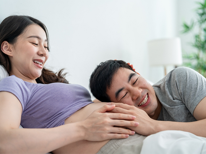 An Asian husband putting his ear to his 14 weeks pregnant wife’s belly in the bedroom.