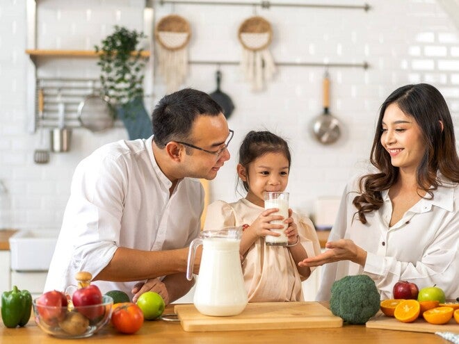 Asian couple watching their little girl drink milk from a glass in the kitchen.
