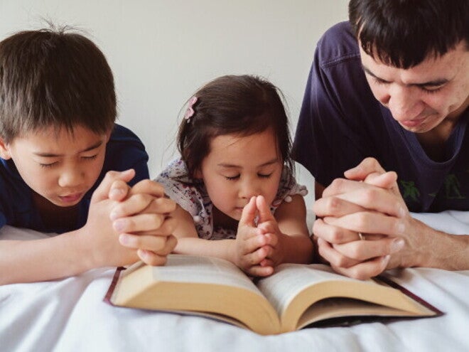 Asian dad with young son and little girl praying on the bed at home, one of their Filipino traditions.