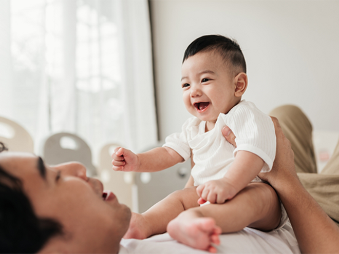 Asian dad lying on bed talks to his smiling baby who's sitting on his stomach