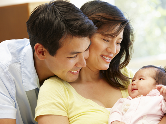 Asian father embraces his wife as she carries their infant who smiles at them, a new step in the child development stages.