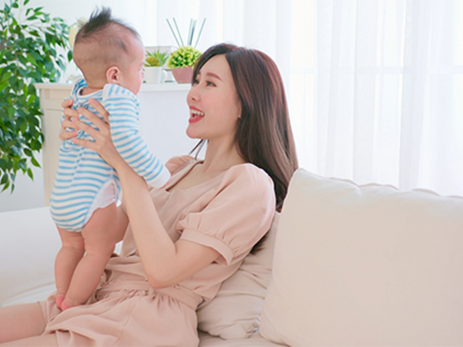 Asian mother holds and talks to her baby on the sofa at home, feeling healthy with proper postpartum care.