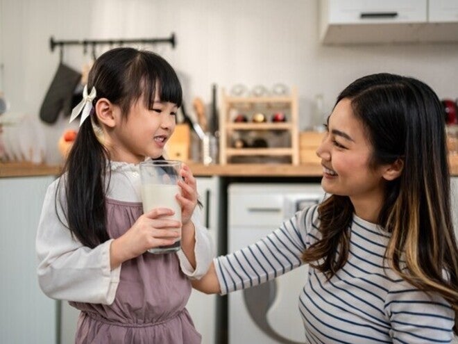 Asian preschooler girl with a glass of milk as her mom smiles at her in the kitchen, happy she’s getting protein in milk