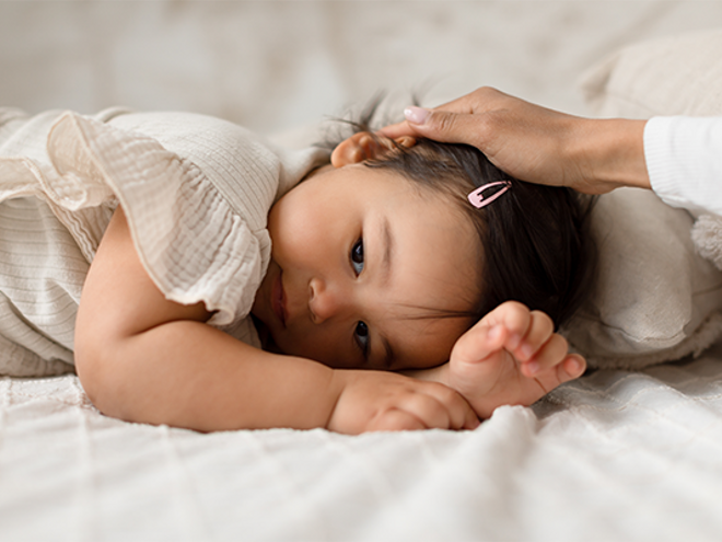 Asian toddler with respiratory tract infection lies face down as her parent strokes her head