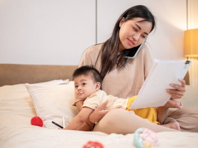 Asian working mom with her baby in bed talks on the phone while looking at documents.