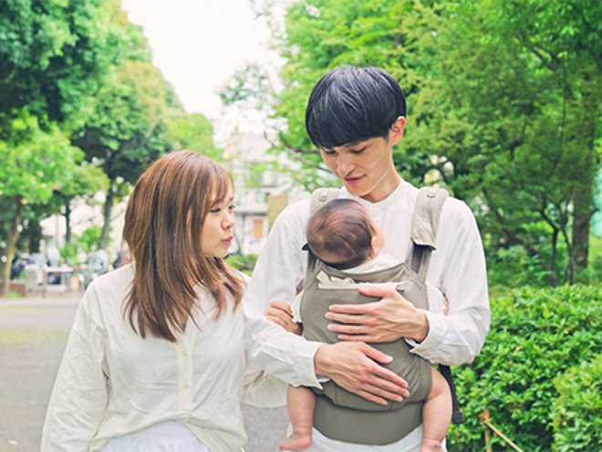 Young Asian dad with wife beside him brings his little one outdoors in a soft-structured baby carrier. Young Asian dad with wife beside him brings his little one outdoors in a soft-structured baby carrier.