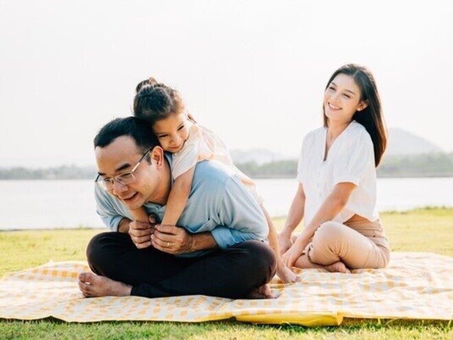 Little Asian girl hugs her dad as they spend an afternoon in the park for Father’s Day, while mom smiles and watches.