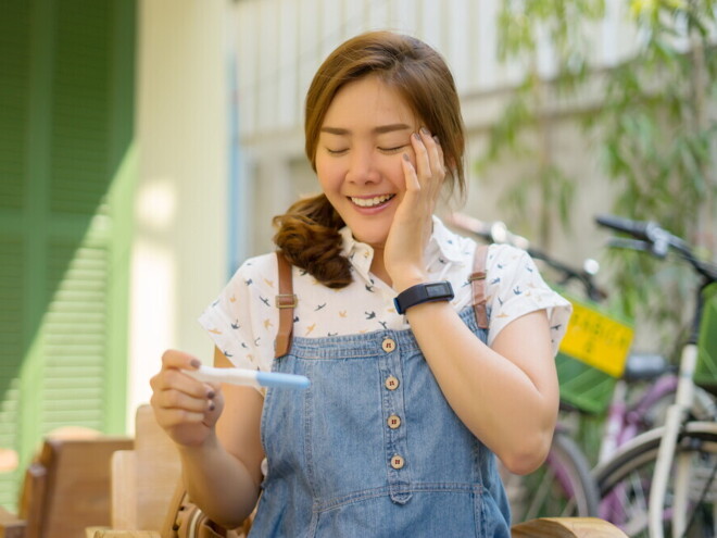 Asian woman in denim jumping smiling at pregnancy test results.