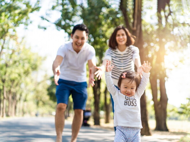 Cute Asian toddler wearing a “girl boss” shirt lifts her arms as her parents reach out to her in the background. Cute Asian toddler wearing a “girl boss” shirt lifts her arms as her parents reach out to her in the background.