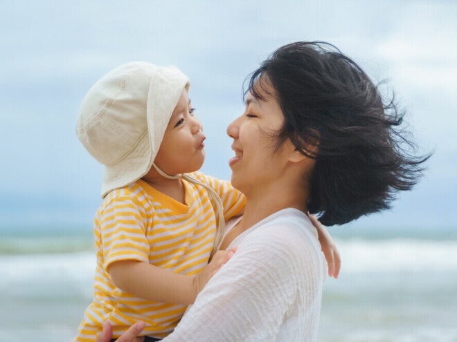 Asian young mom smiles at her talkative toddler son in her arms during a day out in the beach. 