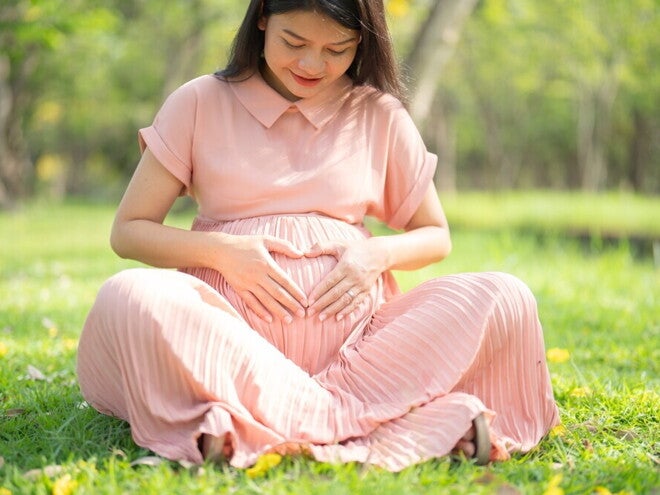 Folic Acid for Pregnancy Asian pregnant woman sitting on grass touches her belly, forming a heart shape with her fingers.