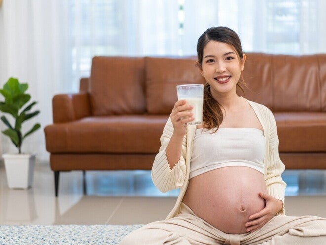 A Practical Guide to Calcium in Pregnancy Pregnant Asian woman holding a glass of milk in front of brown sofa.