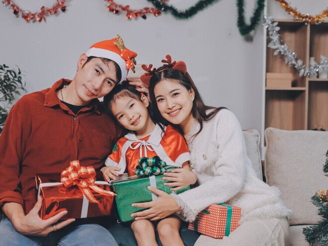 A young Asian couple with their little girl posing for a Christmas photoshoot while holding Christmas presents and wearing holiday headwear.