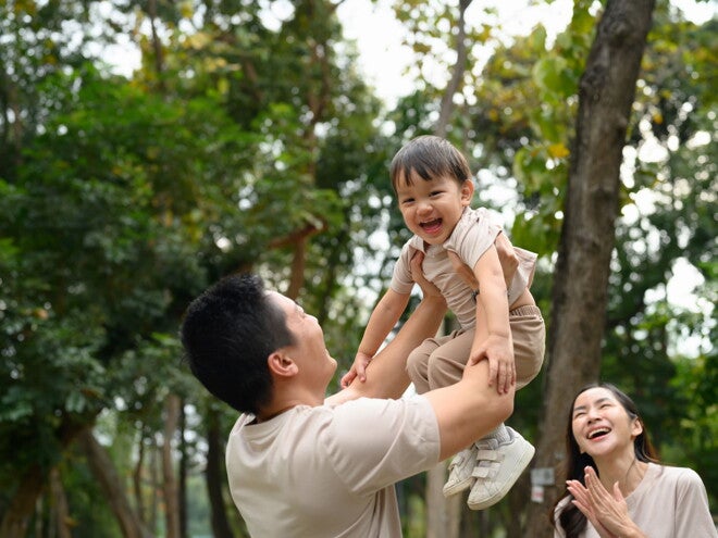 Cropped photo of a dad outdoors lifting his little boy in the air as a form of risky play that builds self-confidence in kids while mom looks on 