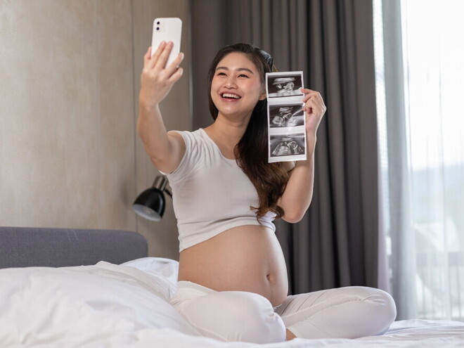 Pregnant Asian takes a selfie in bed while holding her pregnancy ultrasound picture. Pregnant Asian takes a selfie in bed while holding her pregnancy ultrasound picture.
