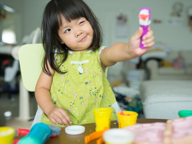 Header_Image Asian preschool-age girl lifts a toy and tilts her head as she looks at it with one eye closed.