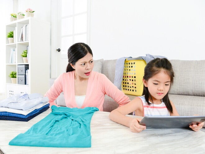 A young Asian mom is getting angry at her little girl playing on a tablet as she folds laundry.