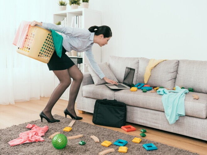 Young Asian mom stressed at messy living room with toys and laundry, trying to tidy up.