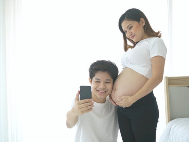 Young Asian pregnant couple taking selfie by a window for bright and beautiful baby bump pictures.