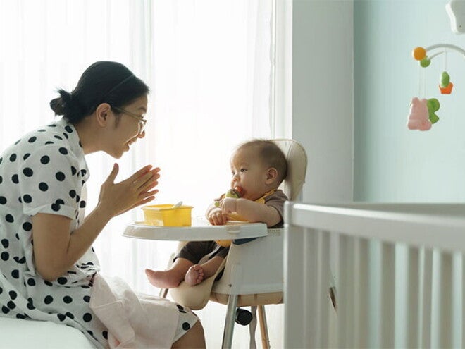 Young Asian mom claps her hands as she watches her baby eating in a high chair.