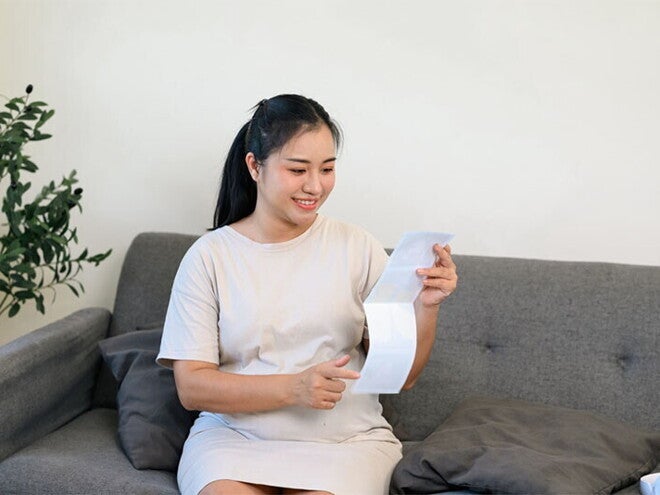 Young Asian pregnant woman on the sofa, holding her tummy and looking at her ultrasound scan at 28 weeks pregnant Young Asian pregnant woman on the sofa, holding her tummy and looking at her ultrasound scan at 28 weeks pregnant