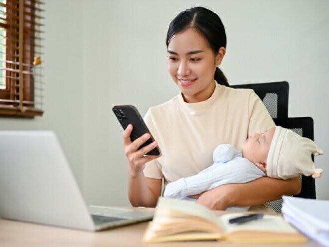 Young freelancing Asian mom checks PhilHealth requirements on her mobile with her baby in her arms.