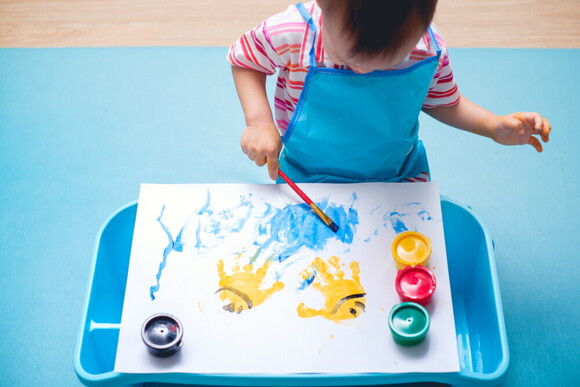 Article_Image-1 Asian toddler uses a brush to paint on paper with yellow handprints.