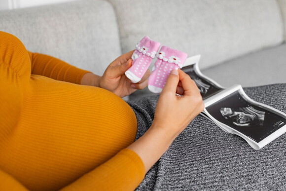Female hands hold a pair of pink socks near baby bump with ultrasound pictures on her lap.