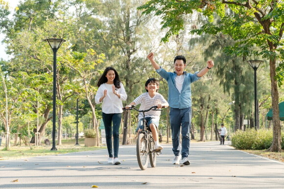 Asian parents cheer on their son as he bikes on his own at a park.