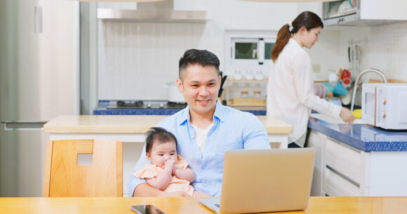 Young Filipino dad works on his laptop with his baby on his lap and his wife working in the kitchen behind him.