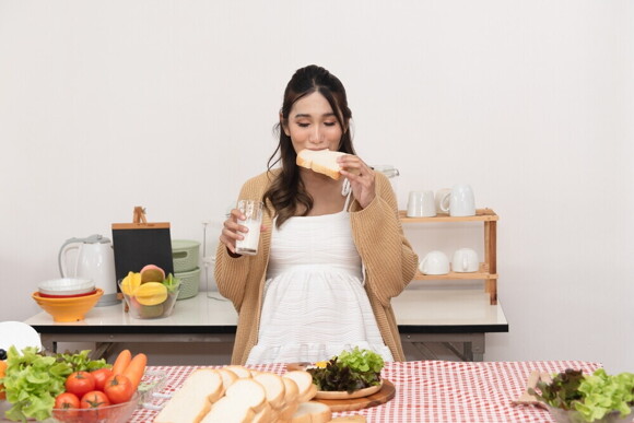One simple rule when it comes to remembering what foods to avoid when pregnant: no raw proteins! Asian pregnant woman in white dress preparing a meal while eating white bread and drinking milk.