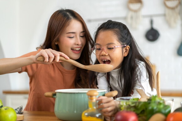 Article_Image-2 Asian mom lifts a ladle from a cooking pot to offer her daughter a taste.