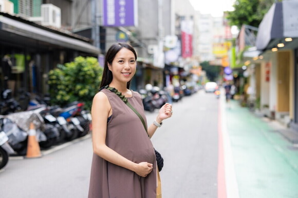 Pregnant Asian holds her belly while walking along a street doing errands.